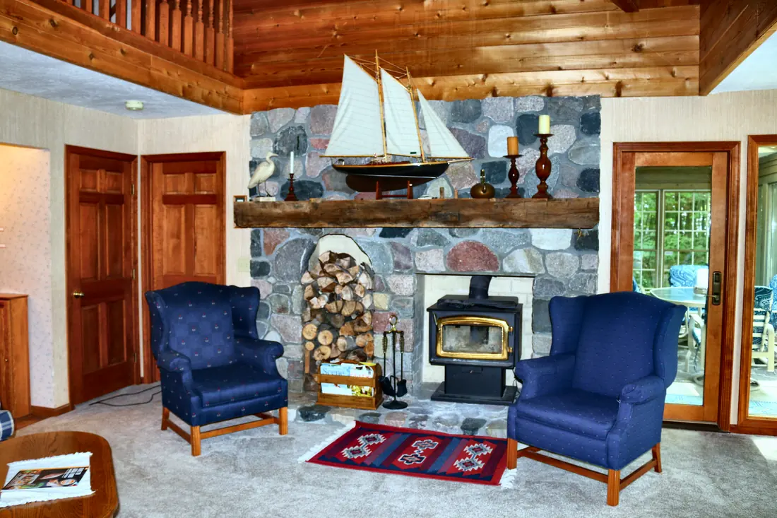 Great room with a fieldstone fireplace, wood stove, timber mantel with a model schooner, and two navy wingback chairs